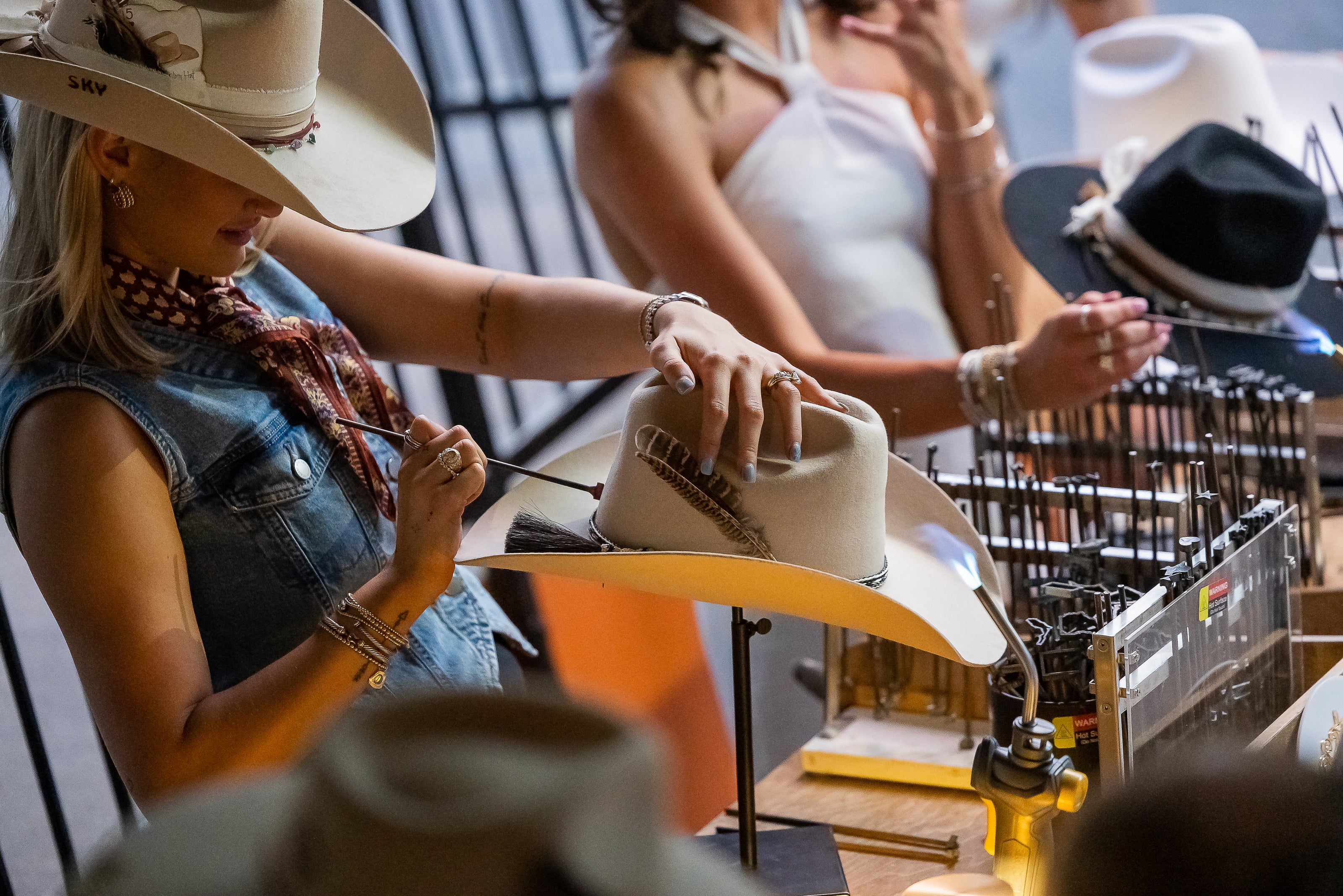 Close-up of a woman shaping and adding a feather to a cream felt cowboy hat with a torch and tools at a busy hat bar