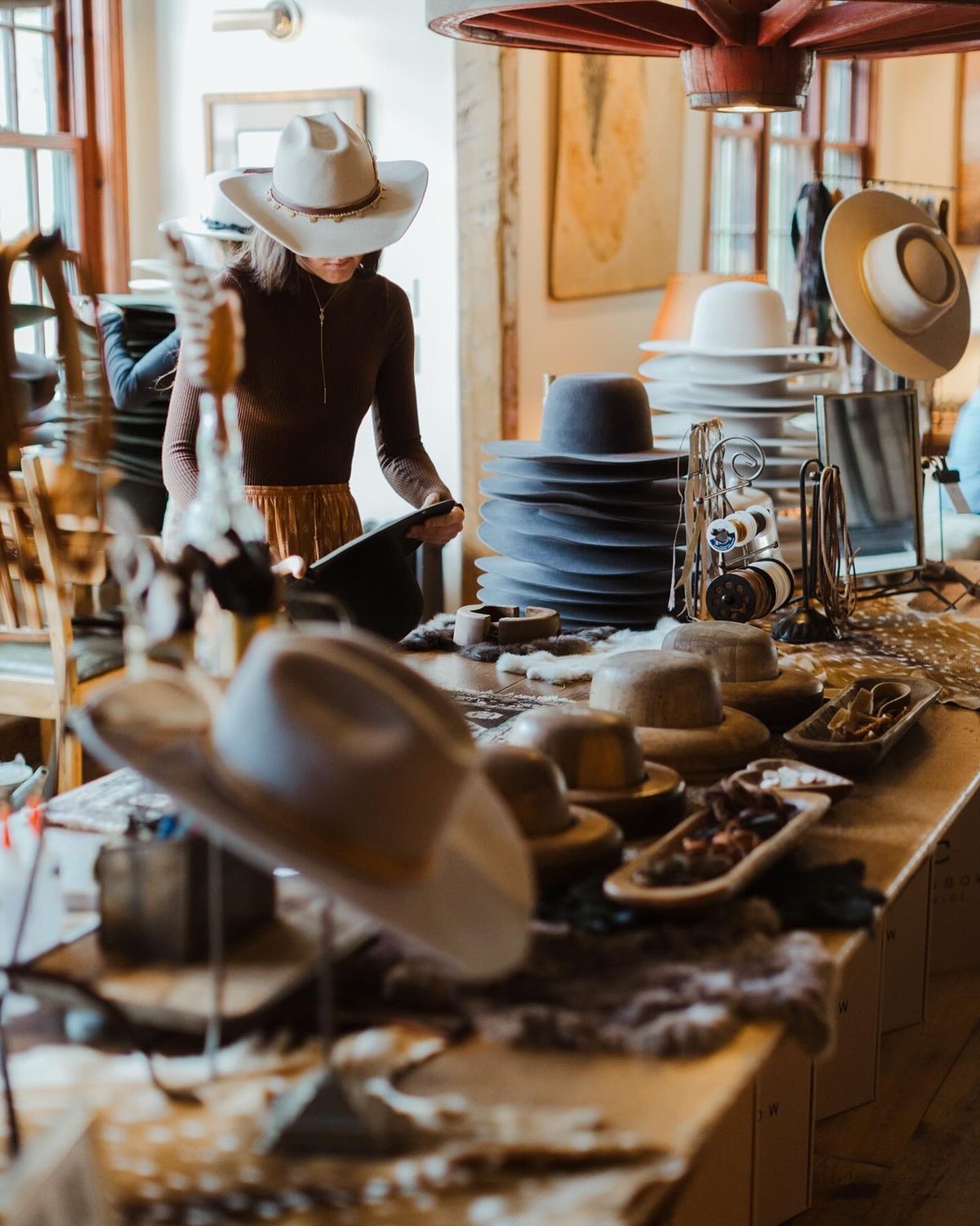 A woman browsing stacks of felt cowboy hats among wooden hat blocks and hides in a warm, rustic hat shop interior