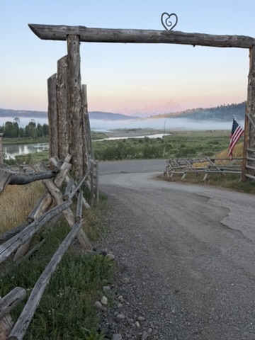 A rustic timber ranch gate with a heart-shaped iron emblem at dawn, fog rising over the valley toward the Grand Tetons, an American flag at the entrance.
