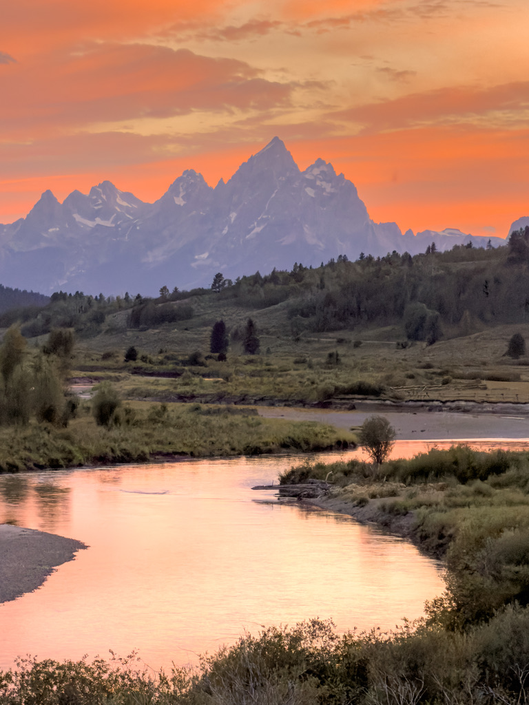 The Grand Tetons at sunset beneath an intense orange sky, purple peaks, with a river in the foreground mirroring the golden light through wetland grasses.