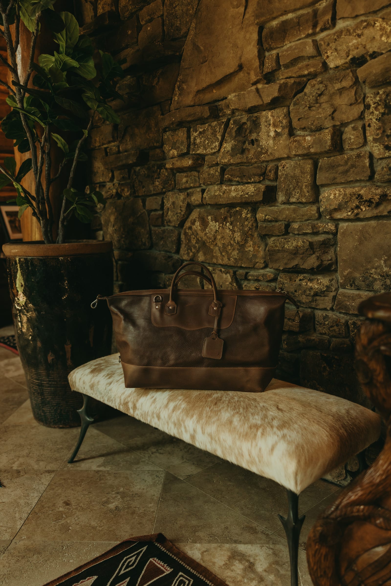 Brown leather travel bag resting on a cowhide bench against a natural stone wall in a Wyoming lodge.