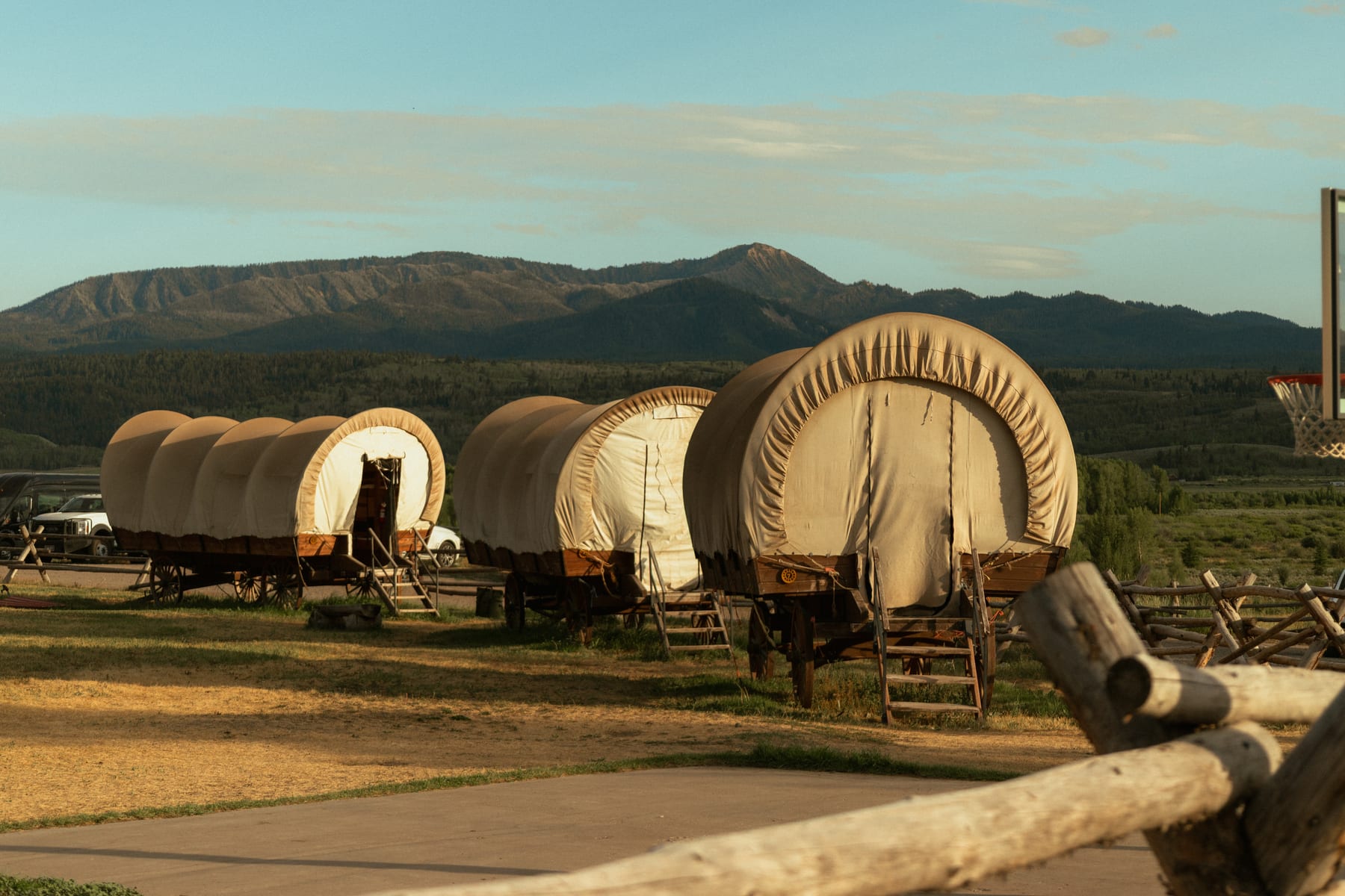 Three canvas covered wagons used as guest lodging at a Wyoming ranch, lit by low morning sun beside a split-rail fence.